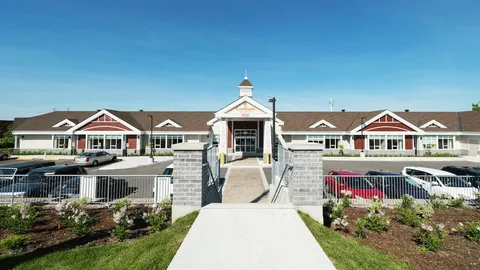 Low-rise building beyond a paved walkway, gardens and parked cars