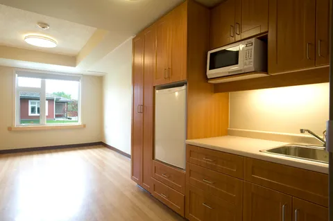 Empty suite with wood flooring, a window and a kitchenette with cabinets and appliances.