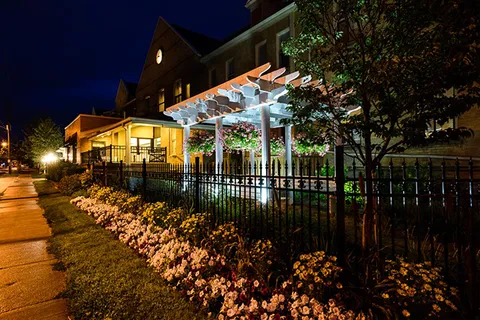 Residence exterior at night beside a white pergola and illuminated flower beds