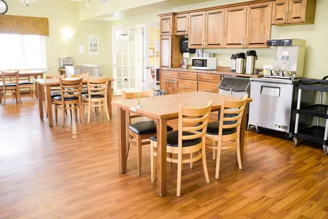 Room with wooden dining tables and chairs beside cupboards, a fridge and drink dispensers