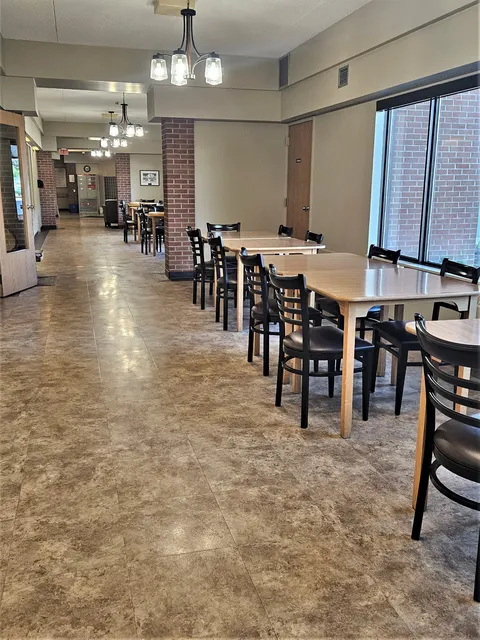 Tables and black chairs along a tiled corridor beside large windows