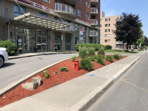 Glass-canopied entrance beside a landscaped driveway and flower beds