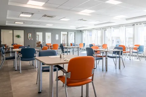 Dining room with orange and blue chairs, square tables and large windows.