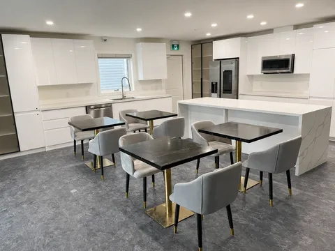 Dining tables and grey chairs beside a white kitchen with a refrigerator and microwave