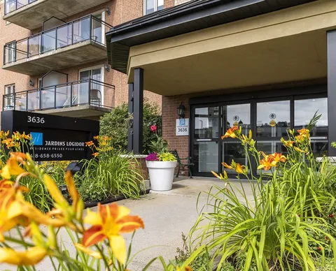 Covered glass entrance beside flower beds and balconies.