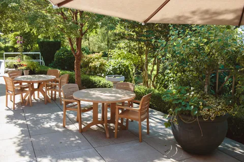 Shaded patio with wood tables and chairs, an umbrella and garden plants.