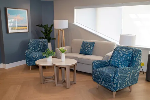 Lounge with a sofa, blue patterned armchairs, round tables and potted plants.