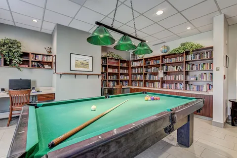 Billiards table beneath green lights beside library shelving.