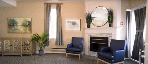 Two blue armchairs beside a fireplace, round mirror, plants and a landscape painting