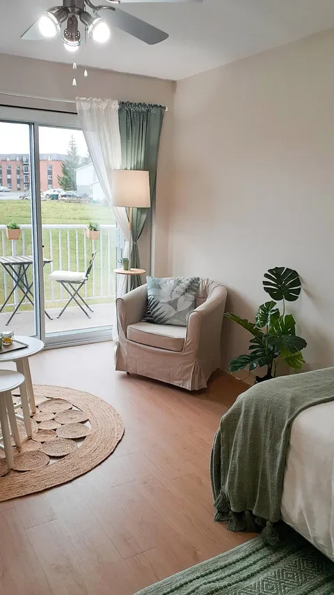 Studio suite with a bed, armchair, plants and a balcony window.