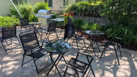 Patio chairs and round glass tables beside a barbecue and planted beds