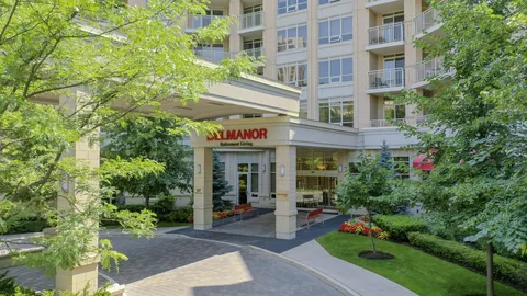 Building entrance beneath balconies, with trees, flower beds and a curved driveway