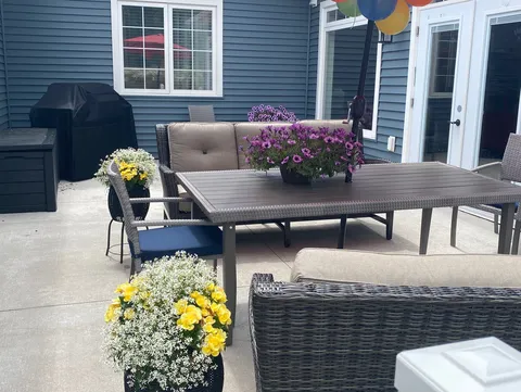 Patio with wicker chairs, a rectangular table, a covered barbecue and pots of flowering plants