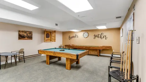 Room with a pool table, shuffleboard table, chairs and a cue rack