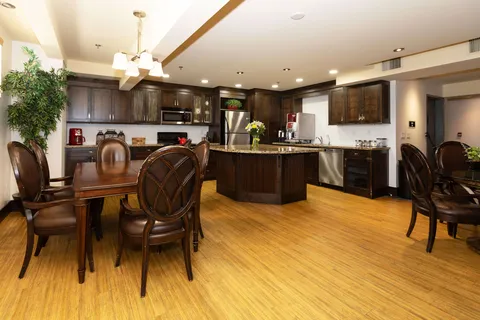 Table and upholstered chairs beside a kitchen with dark cabinets and stainless-steel appliances