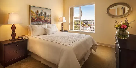 Bedroom with white bedding, two lamps, a dresser and a city view through a window