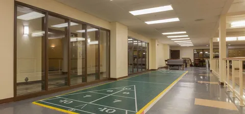 Indoor shuffleboard court with seating beyond and glass-walled rooms beside it
