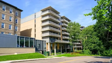 Multi-storey yellow and grey residential building with balconies, lawn and trees beside a road