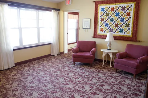 Two purple armchairs beneath a colourful quilt beside a small table and a lamp