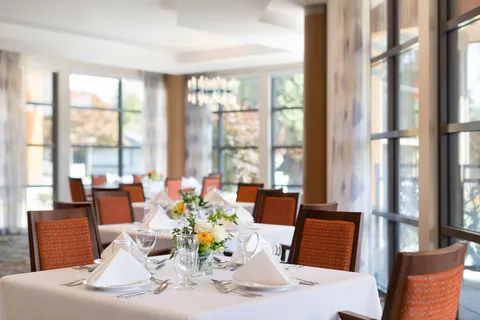 Dining room tables beside tall windows, set with flowers and glassware.