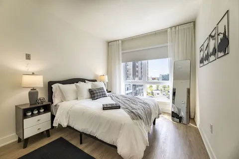 Bedroom with a white bed, bedside lamp and a window overlooking nearby buildings
