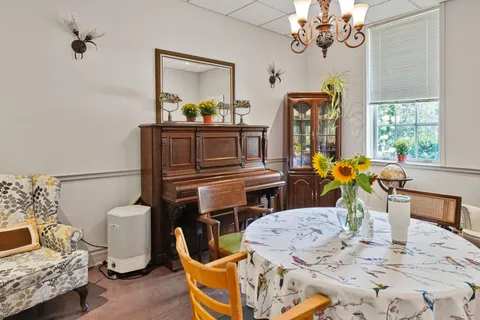 Room with an upright piano, wooden chairs, a round table and a glass-front cabinet