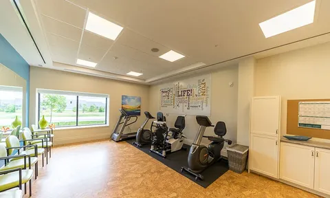 Fitness room with exercise machines and chairs beside windows.