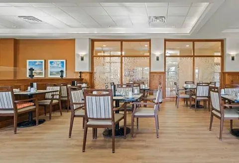 Dining room with tables and wooden chairs beside large windows.