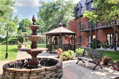 Garden with a tiered fountain, gazebo, metal bench and flowering plants beside the brick building