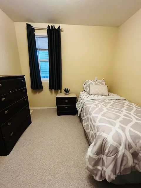 Bedroom with a patterned bedspread, dark chest of drawers and black curtains.
