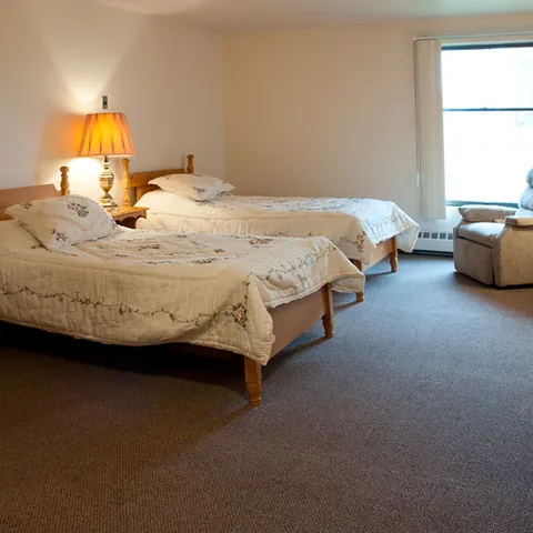 Bedroom with two wooden beds, floral bedding, a bedside lamp and an armchair by a window