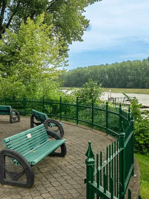 Paved riverside seating area with green benches and a metal fence.