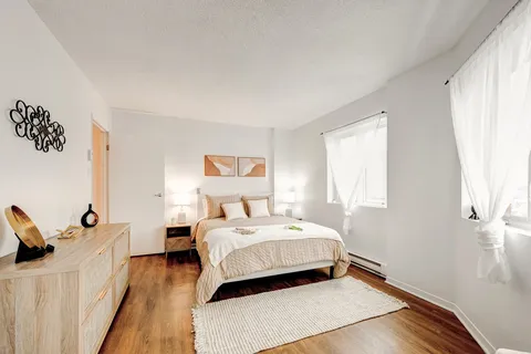 Bedroom with pale bedding, wooden dresser, bedside lamps and curtained windows