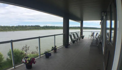 Covered riverside balcony with outdoor chairs and potted flowers.