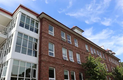 Brick residence facade with white bay windows, trees and red roof trim under a blue sky