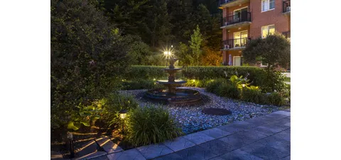 Garden fountain and planting beds lit beside a paved path at dusk
