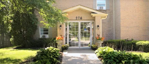 Glass entrance doors beneath a small canopy, with flower planters and surrounding greenery