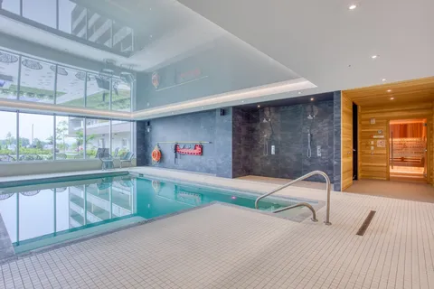 Indoor swimming pool beside tall windows and a sauna entrance.