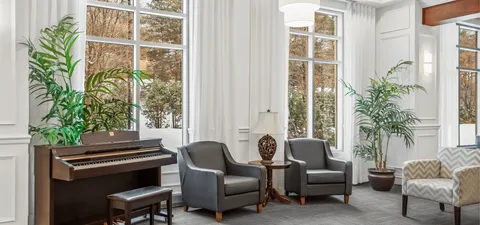 Upright piano and armchairs beside tall windows and potted plants