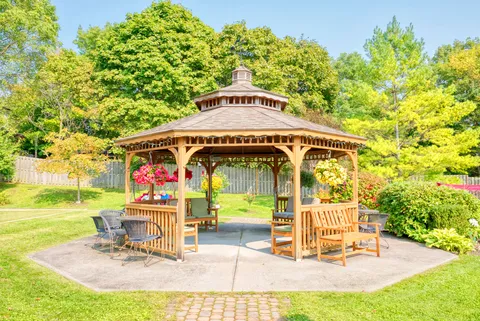 Wooden gazebo with benches and flower baskets beside trees and a lawn