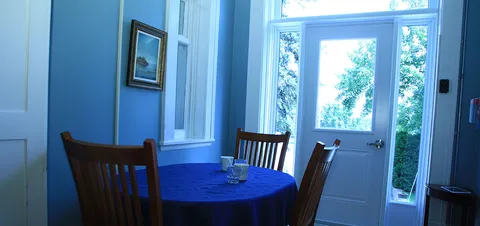 Blue dining room with wooden chairs, a round table and windows beside a door.