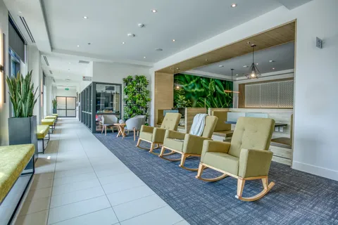 Common room with green rocking chairs, tables and a leafy wall mural.