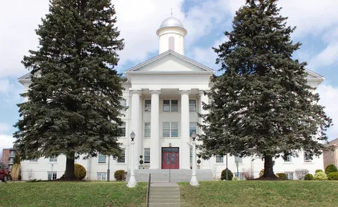 White building with tall columns, a cupola, red doors and evergreen trees