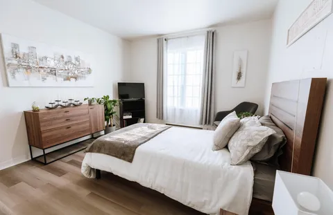 Bedroom with a white bed, wooden dresser, black chair and curtained window