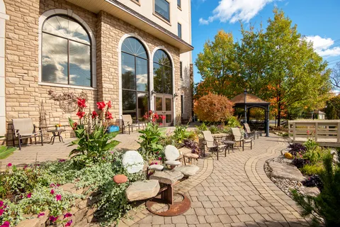 Patio chairs, flower beds, a gazebo and a small bridge beside arched residence windows
