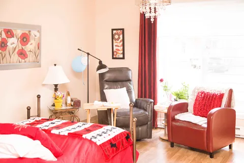 Bedroom with red bedding, two armchairs, floral artwork and a large window