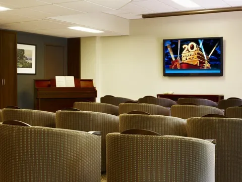 Room with rows of upholstered chairs facing a television beside a piano