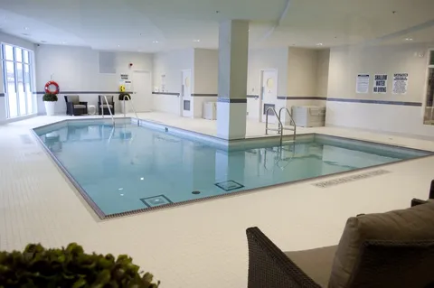 Indoor swimming pool with handrails and seating beside large windows.