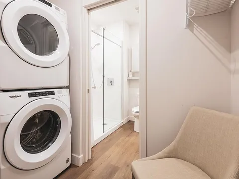 Stacked washer and dryer beside a chair and an open bathroom doorway