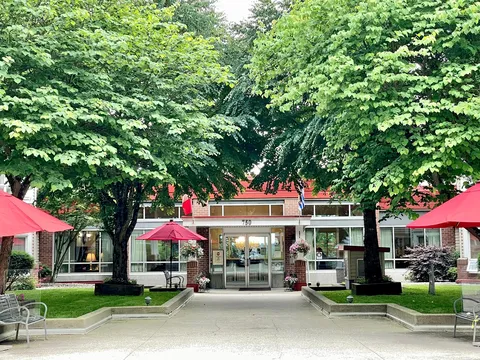 Courtyard path leading to glass entrance doors beneath trees and red umbrellas.